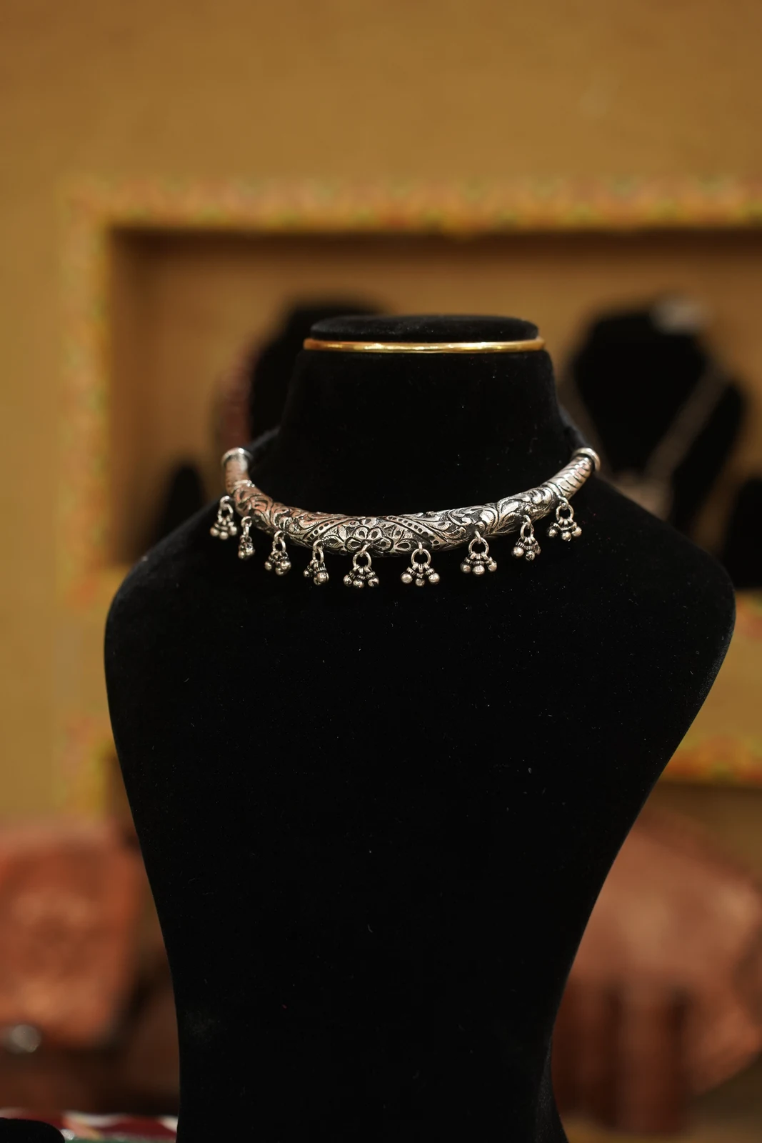 Silver-coloured contemporary Hansuli collar with scrolling decoration and small hanging clusters, shown on a black display bust.