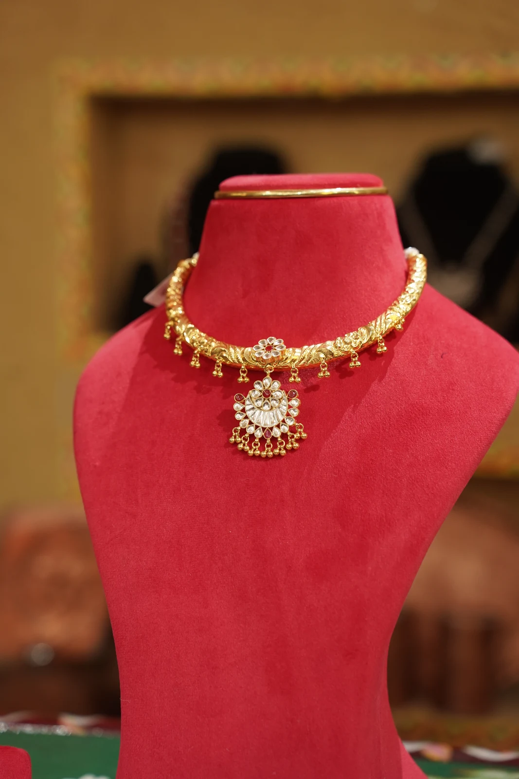 Gold-coloured contemporary Hansuli collar with a central floral pendant and hanging clusters, shown on a red display bust.